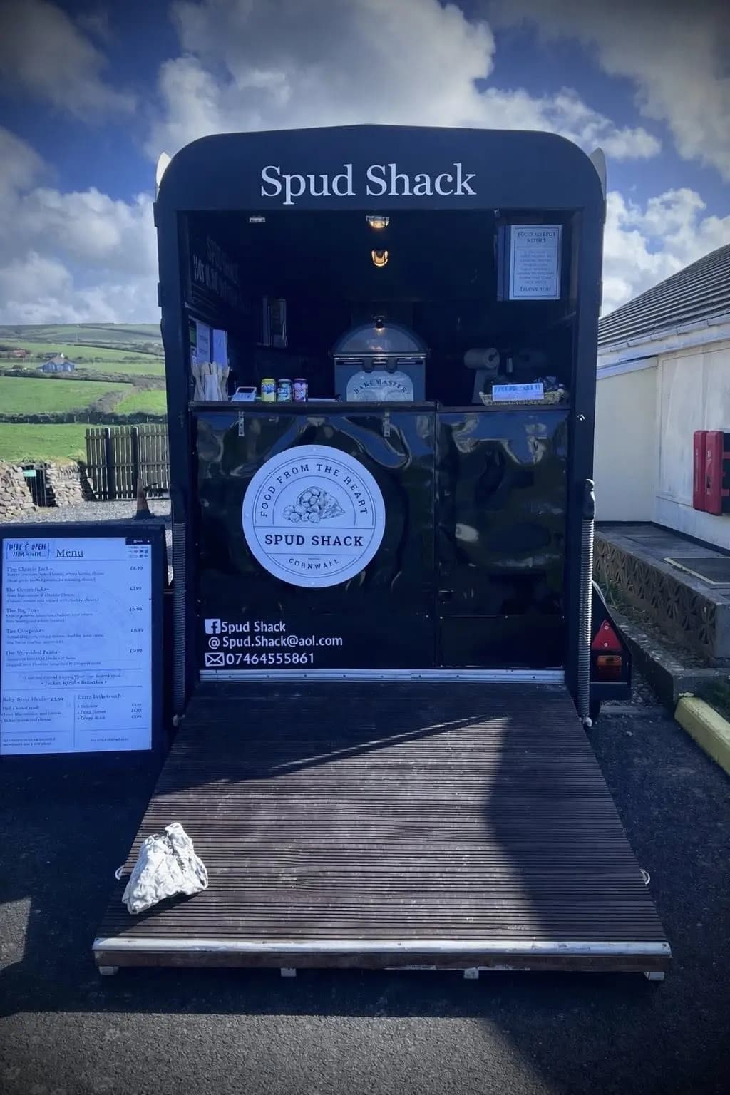 The Spud Shack horsebox open and trading — front view of the trailer with the Spud Shack sign overhead and the Food From The Heart roundel below, menu board on the left, Cornish hills behind